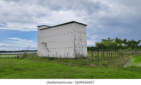The image displays a tall, white rectangular building with numerous small circular openings, presumably a bird's nest farm, standing in a green field under a partly cloudy sky.
