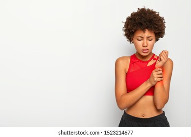 Image Of Displeased Afro Girl Hurt Arm During Workout, Touches Wrist, Suffers From Painful Feelings, Needs To See Surgeon, Wears Casual Outfit, Poses Against White Background, Copy Space For Advert