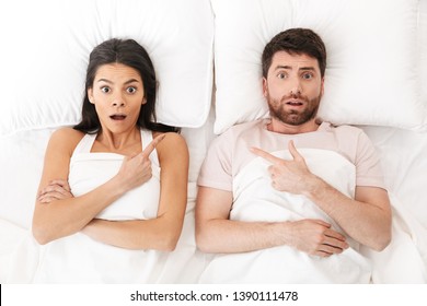 Image Of A Displeased Shocked Scared Confused Young Loving Couple In Bed Under Blanket Pointing.