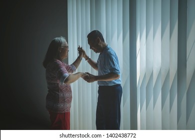 Image Of Elderly Couple Looks Happy While Dancing Near The Window. Shot In The Dark Room