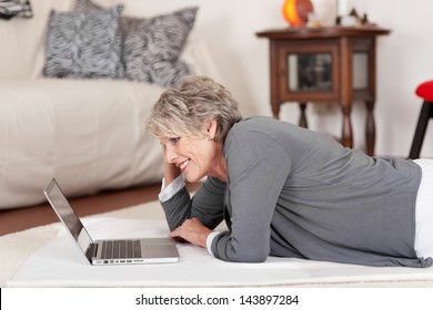 Image of an elderly happy woman lying on the floor and working on the laptop.