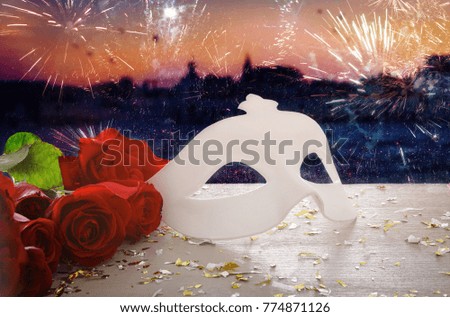 Image of elegant venetian mask and red roses over wooden table in front of blurry Venice background