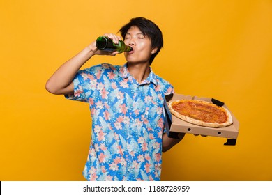 Image Of Emotional Excited Young Asian Man Standing Isolated Over Yellow Background Holding Pizza Drinking Beer.