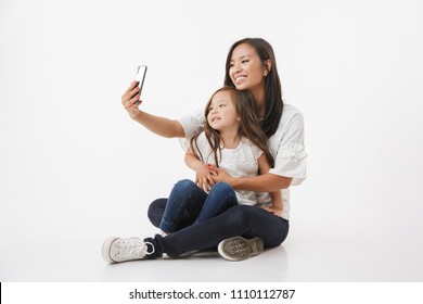 Image Of Emotional Happy Young Asian Woman Mother With Her Little Girl Child Daughter Sitting Isolated Over White Wall Background Make Selfie By Mobile Phone.