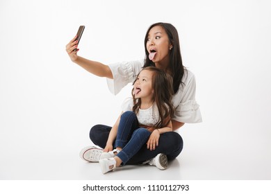 Image Of Emotional Happy Young Asian Woman Mother With Her Little Girl Child Daughter Sitting Isolated Over White Wall Background Make Selfie By Mobile Phone.