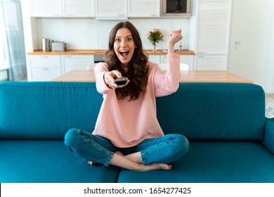 Image Of Emotional Smiling Optimistic Young Woman Sit Indoors At Home Watch Tv Holding Remote Control On Sofa Make Winer Gesture.