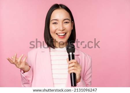 Image of enthusiastic asian businesswoman giving speech, talking with microphone, holding mic, standing in suit against pink studio background