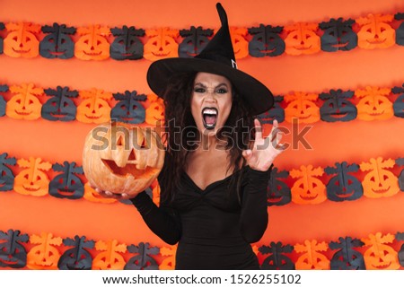 Image of evil witch girl in black halloween costume holding carved pumpkin isolated over orange wall
