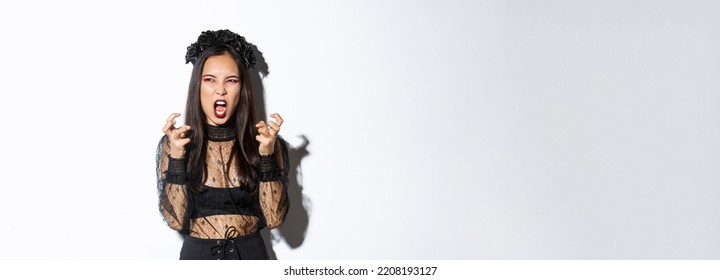 Image of evil witch hate something, looking angry and furious at upper left corner, cursing or swearing while clenching fists mad, standing in halloween costume over white background - Shutterstock ID 2208193127
