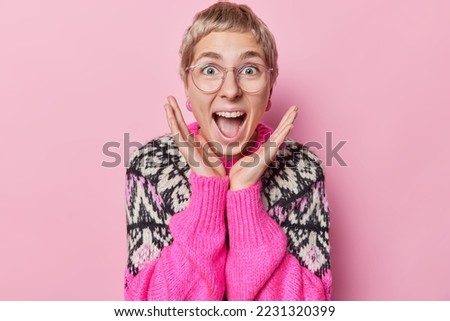 Image of excited beautiful woman with short hair raises hands over face yells loudly keeps mouth widely opened reacts to something awesome wears big spectacles and warm jumper. Wow thats great