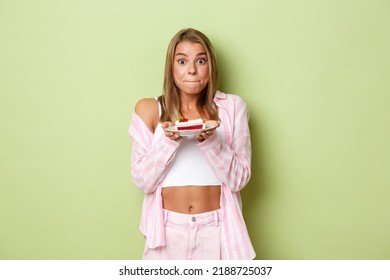 Image Of Excited Blond Girl Holding Piece Of Cake, Tempted To Taste Dessert, Standing Over Green Background