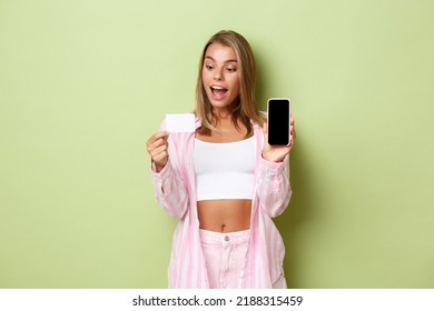 Image Of Excited Blond Girl In Pink Shirt, Showing Credit Card And Mobile Phone Screen, Standing Over Green Background