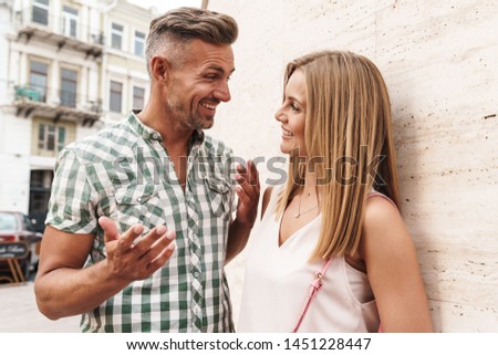 Image of excited blonde couple in summer clothes smiling and talking together while standing against wall on city street