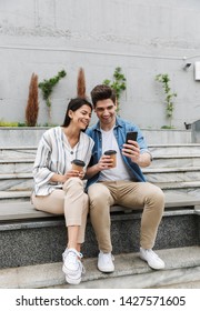 Image of excited couple man and woman in casual clothes drinking takeaway coffee and using smartphone on city stairs outdoors