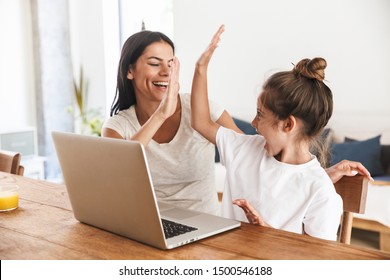 Image of excited family woman and her little daughter giving high five and using laptop computer together while sitting at table in apartment