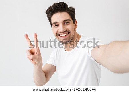 Image of excited happy young man posing isolated over white wall background make a selfie by camera showing peace gesture.