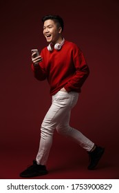 Image Of Excited Young Asian Man Using Headphones And Cellphone While Walking Isolated Over Burgundy Background
