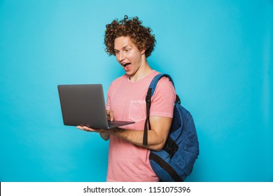 Image Of Excited Young Guy With Curly Hair Wearing Casual Clothing And Backpack Holding And Using Open Laptop Isolated Over Blue Background