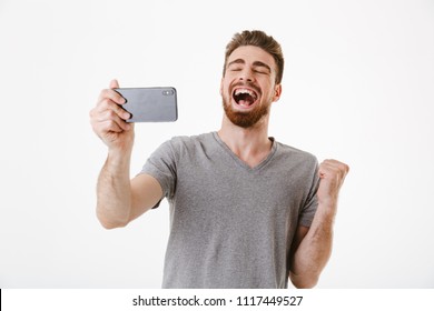 Image Of Excited Young Man Standing Isolated Over White Wall Background Play Games By Mobile Phone Make Winner Gesture.