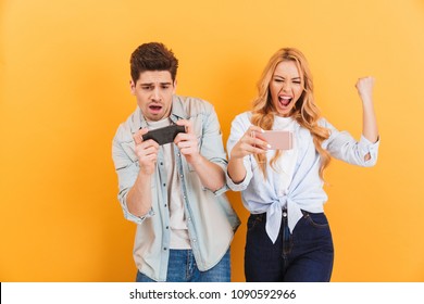 Image of excited young man and woman playing together and competing in video games on smartphones isolated over yellow background