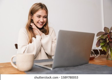 Image of excited young woman wearing casual clothes using laptop and drinking tea in cozy room