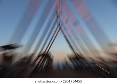 The image features an abstract, dynamic perspective of a triangular or A-frame wooden architectural structure, captured with an intentional zoom burst or motion blur technique during a serene dusk or dawn. The camera movement creates dramatic, exploding lines that radiate outward from the central apex, giving a powerful sense of speed, energy, and hyper-lapse motion. The warm, earthy tones of the wooden beams blend softly with the gradients of the twilight sky, transitioning from a clear, tranquil blue at the top to a subtle orange and pale yellow horizon line at the base. The blurred background hints at an outdoor coastal or beach setting with dark silhouetted trees or structures, making this conceptual photograph ideal for themes related to architectural design, fast-paced modern life, abstract transitions, and creative long-exposure photography.