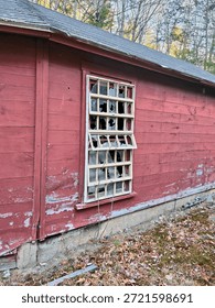 The image features a broken window on a red building. The structure appears to be an abandoned shed or barn, made of wooden materials that are painted red. The surrounding ground shows signs of decay, emphasizing the neglected state of the property.