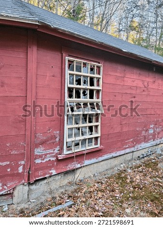 The image features a broken window on a red building. The structure appears to be an abandoned shed or barn, made of wooden materials that are painted red. The surrounding ground shows signs of decay, emphasizing the neglected state of the property.