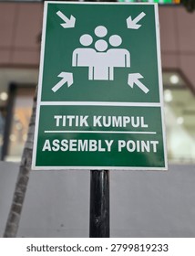 The image features a close-up, eye-level shot of an emergency evacuation or gathering sign mounted on a black metal post.
​The sign is rectangular and has a dark green background with a thin white border. It is divided into two main sections:
​Top Section: This part displays a standard pictogram of four stylized human figures standing closely together. Four white arrows, located in each corner of this section, point diagonally inward toward the figures to denote a gathering center.
​Bottom Section: This section contains bilingual text in bold, white, capital letters. which is underlined by a thin white line. Directly underneath it, the English translation reads ASSEMBLY POINT.
​The Post: The sign is securely attached to a vertical black metal pipe or pole at the bottom center.
​The Background: The background is softly blurred shallow depth of field. It shows the exterior of a modern building with large glass windows or storefronts, light-colored walls, and the trunk of a slender tree or palm on the left side, indicating an outdoor urban setting.