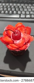 The image features a close-up, high-angle shot of a beautifully decorated cupcake resting on a dark, matte surface. In the background, a slightly blurred computer keyboard is visible, suggesting the photo was taken at a desk or workspace.