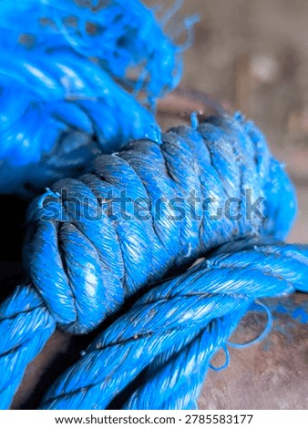 The image features a close-up macro view of thick, vibrant blue synthetic ropes tied into a large, secure knot. The main focus is on the tight coils and twists of the braided strands, which show signs of heavy use, including frayed fibers, dust particles, and slight weathering on the surface. Another layer of the blue rope passes horizontally underneath the main knot, resting against a dark, weathered structure. The background is softly blurred, keeping the emphasis on the detailed texture and vivid color of the cord