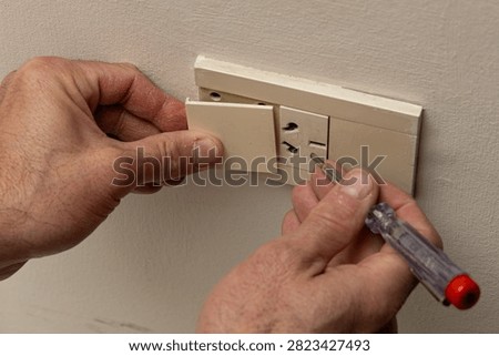 The image features a hand inserting a device into an electrical outlet, which is being repaired with a polarity tester. A person, whose hands are visible on the left side of the frame, appears to be taking care of the task at hand. This content can be described as 'Editorial Illustrations, Vectors and Animations' as it illustrates an event that requires credentials. The description 'A Latin American man's hands taking apart and repairing an electrical outlet in his home using a polarity tester' is accurate to the scene depicted.