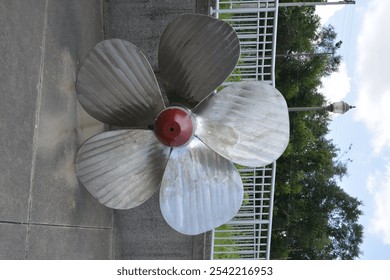 The image features a large, metallic propeller with four blades, prominently displayed outdoors. Its polished surface reflects light, showcasing its design as a crucial component for marine propulsion