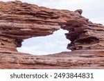 The image features a large rock formation with a natural arch, showcasing textured red sandstone under a cloudy sky. The unique geology creates a striking natural frame in the landscape.