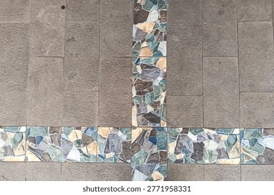 The image features a top-down view of stone tile flooring with a unique decorative cross-pattern. The primary surface consists of rectangular, textured tiles in a neutral grayish-brown earth tone. Intersecting these are bands of mosaic-style tiles, composed of irregular stone fragments in varying shades of blue, green, tan, and dark brown. The layout creates a clean, architectural aesthetic, blending natural textures with a structured geometric design, typical of outdoor patios or modern pedestrian walkways.
