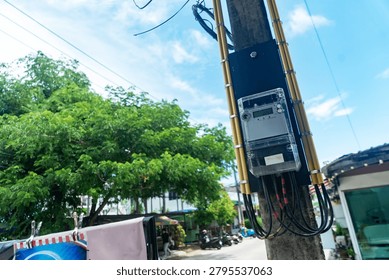 The image features a utility pole equipped with electrical wires and communication cables set against a backdrop of trees and a street scene on a bright, partly cloudy day.