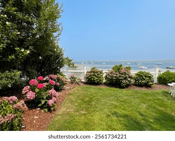 The image features vibrant pink hydrangeas in a well-kept garden, bordered by a white picket fence with boats floating in calm waters beyond under a clear, bright sky.