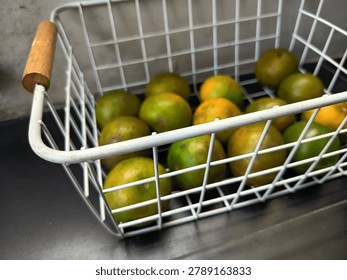 The image features a white wire storage basket with wooden handles, filled with several green and yellowish oranges.