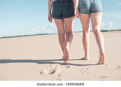 Image of female feet close-up on a beach background. Two women in shorts walk barefoot on the sand, leaving footprints.