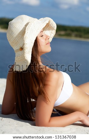 Image of female in white bikini and hat sunbathing on sandy beach during vacation