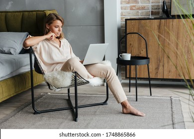 Image Of Focused Blonde Nice Woman In Stylish Pajamas Working With Laptop While Sitting On Armchair At Living Room