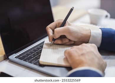 The image focuses on a man's hand scribbling in a notepad on a stylish laptop, set against a blurry scene - Powered by Shutterstock