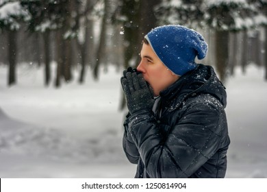 Image Of Freezing Young Man Wearing Black Leather Winter Jacket And Blue Hat Is Breathing Into Hands In Gloves And Warming Himself Standing Among The Cold Snowy Forest.