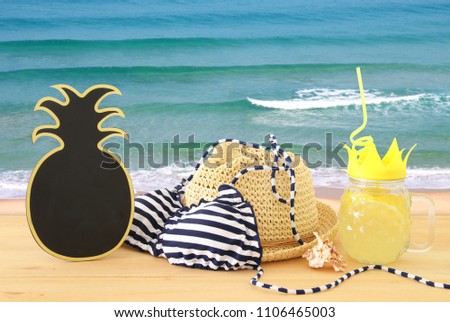Image of fresh lemonade drink in cute pineapple shape glass with twisted straw next to bikini swimsuit and beach fedora hat over wooden table