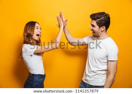 Image of friendly young people man and woman in basic clothing laughing and giving high five isolated over yellow background