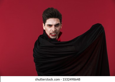 Image Of Frightening Vampire Man With Blood And Fangs In Black Halloween Costume Holding His Cloak Isolated Over Red Wall