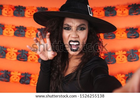 Image of frightening witch girl in black halloween costume screaming and scratching isolated over orange pumpkin wall