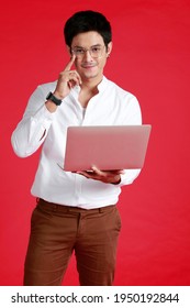 Image Front View  Young Asian Confident Handsome Man Looking To Camera And Working Online With Digital Computer Laptop  While Standing Against Isolated Red Background. Bangkok Thailand South East Asia