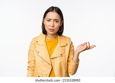 Image of frustrated korean woman, shrugging shoulders and raising hand, looking puzzled, cant understand smth, standing over white background