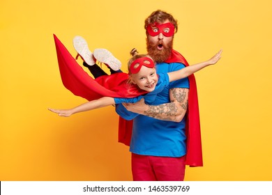 Image of ginger father and daughter dressed in superheroes, surprised man carries little kid on hands, pretend flying and saving world, pose against yellow background. Childhood and fun concept